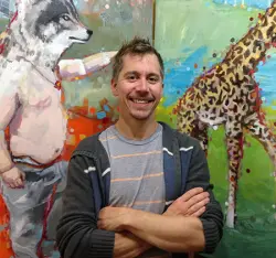 Artist Adam Swanson smiling in front of painting of man in jeans with wolf head and giraffe
